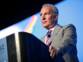 Peter Schiff speaking to the audience behind a desk, wearing a suit and a purple tie.