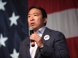 Andrew Yang standing in front of the US flag, with a microphone.